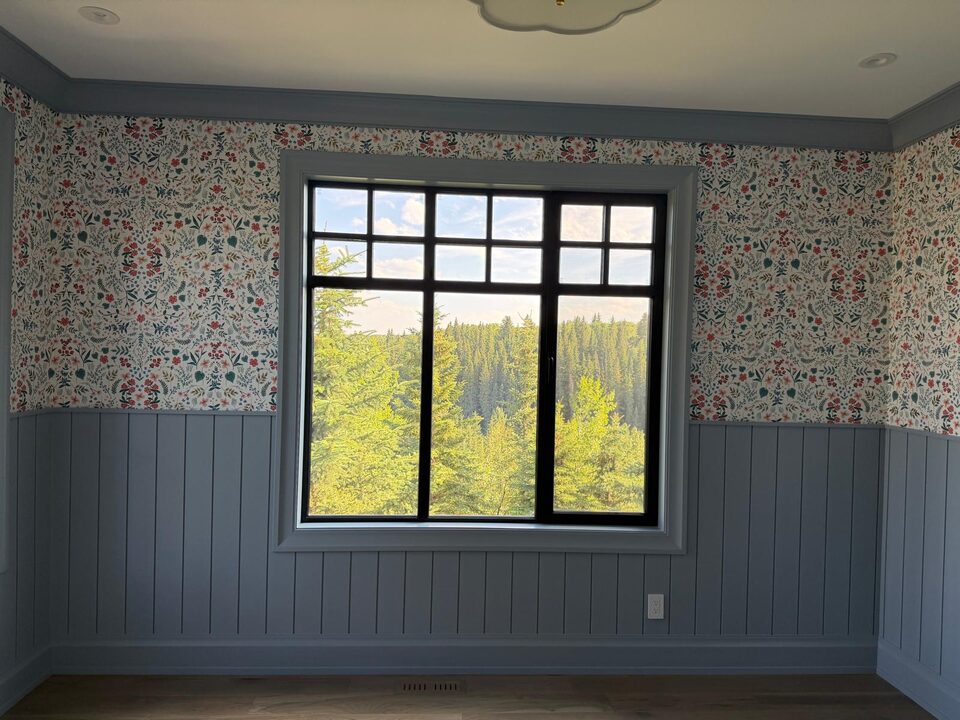 Wildflower wallpaper mirror-matched symmetrically around a large black-frame picture window over a forested valley in an Elbow Valley home near Calgary