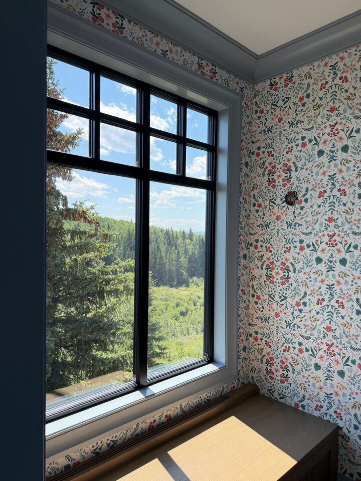 Botanical print wallpaper trimmed around a black-frame window overlooking evergreen forest in an Elbow Valley new build