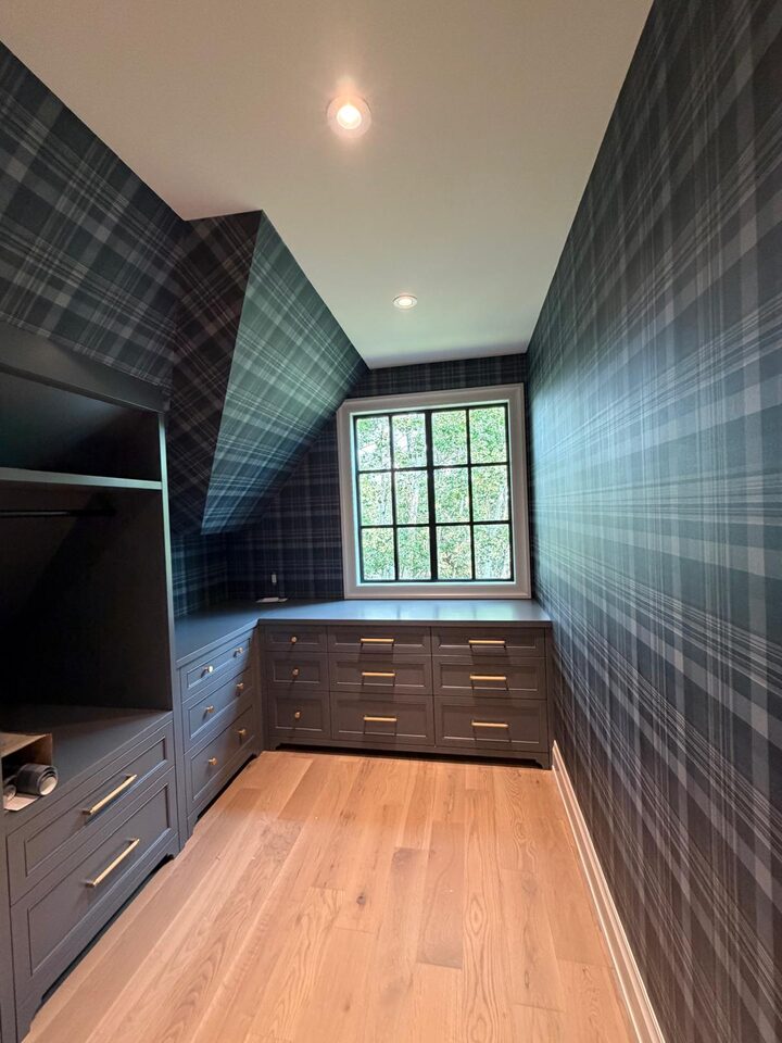 Navy tartan plaid wallpaper wrapping a dressing room around a black-frame window and gray built-ins, matched into the dormer ceilings in an Elbow Valley home west of Calgary