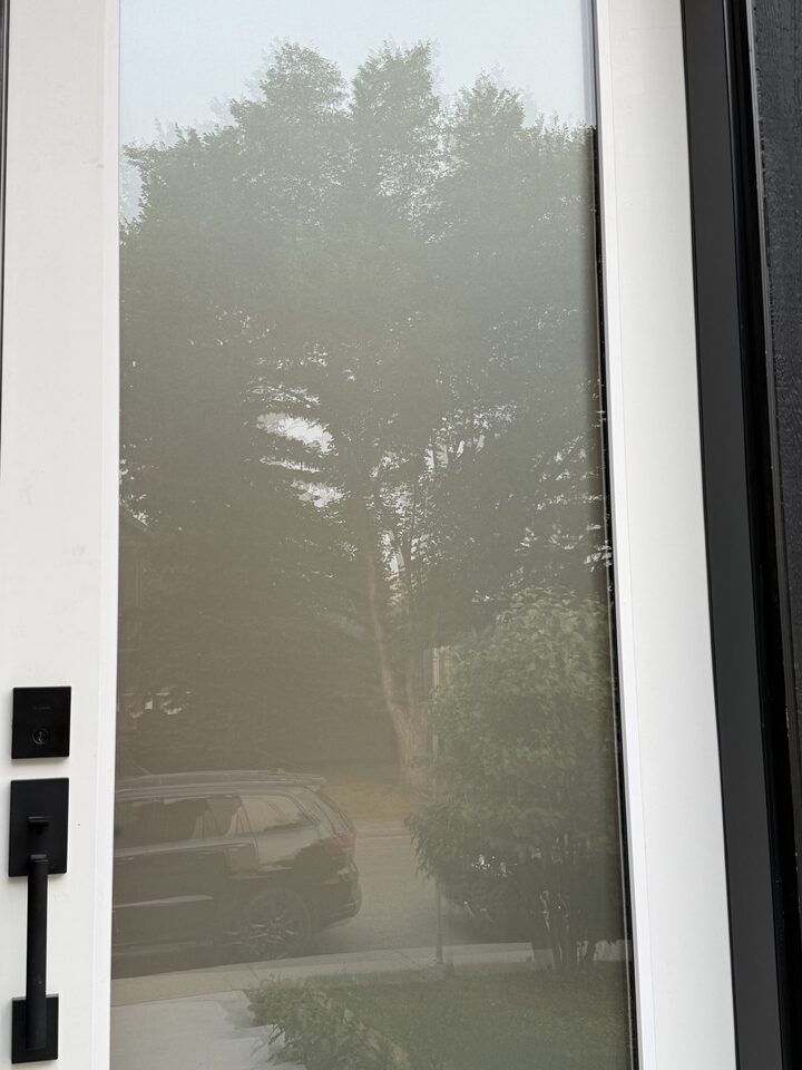 Clear full-lite front door glass before privacy film, photographed from the step outside, the yard and parked vehicle reflecting off the pane, Glenbrook SW Calgary